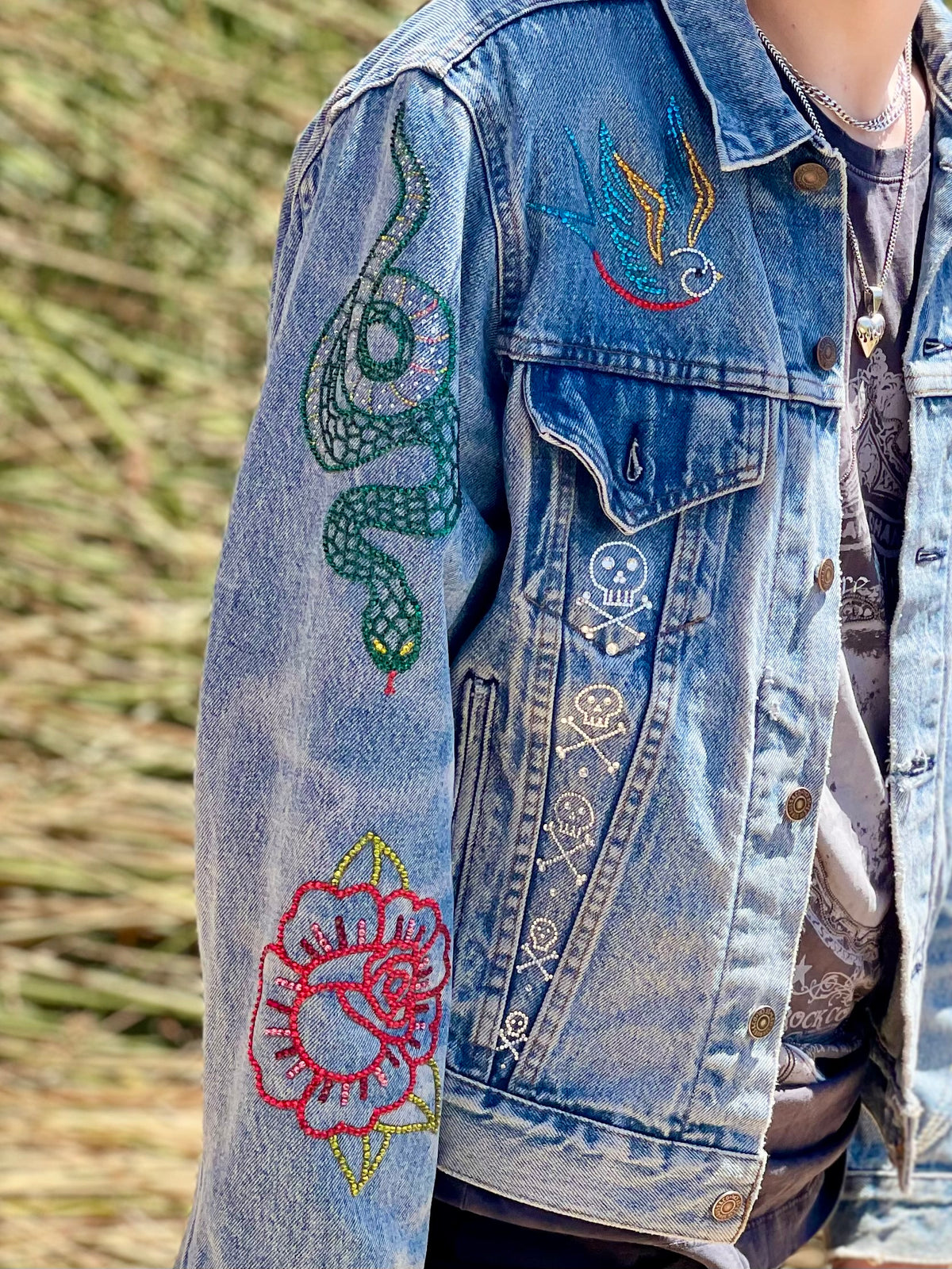 Denim jacket with custom Looking Glass Gems rhinestone embellished designs in classic American tattoo flash style on a blurred natural background