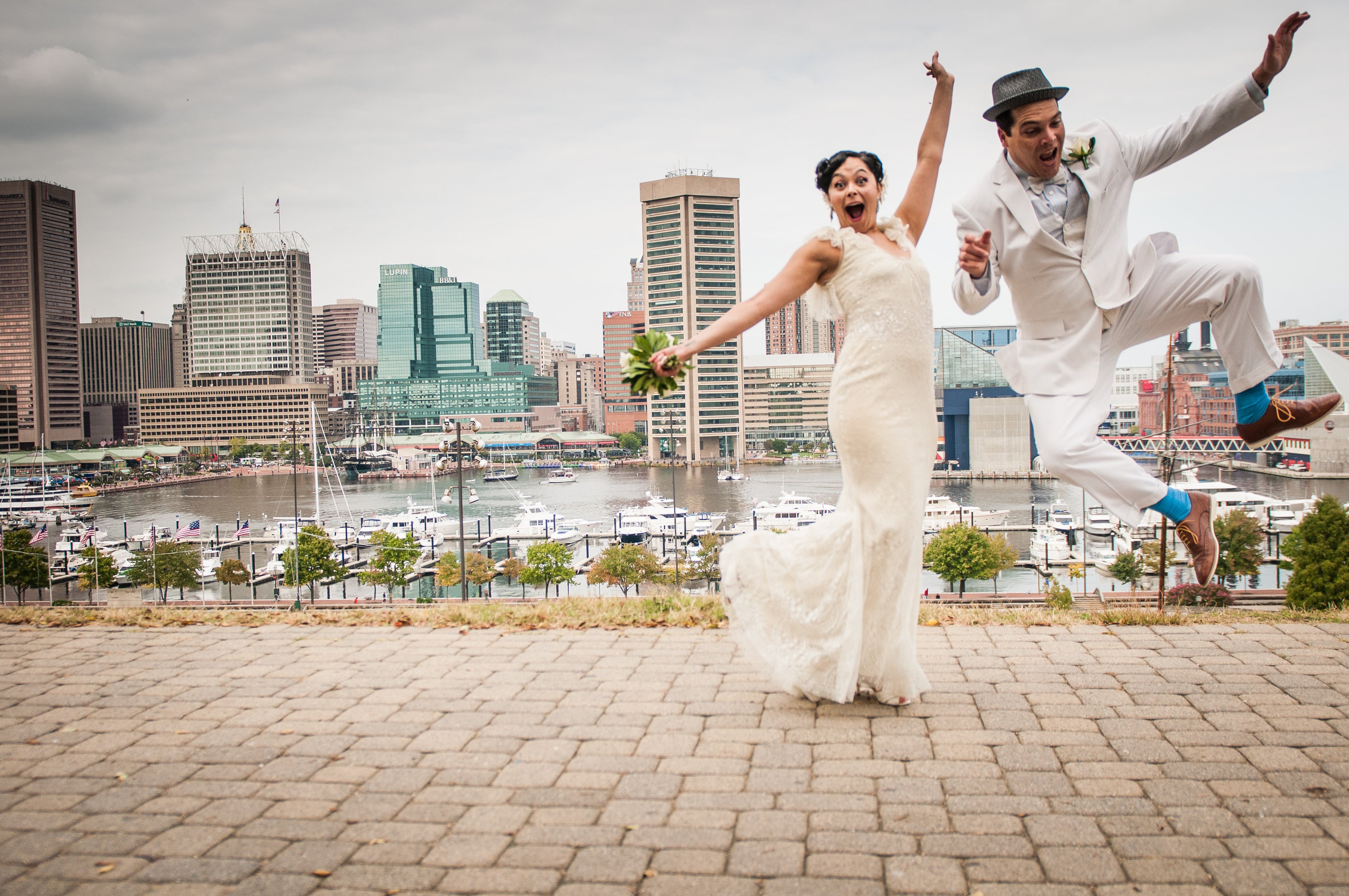 Wedding couple jumping in front of a Baltimore city skyline with boats on the water.