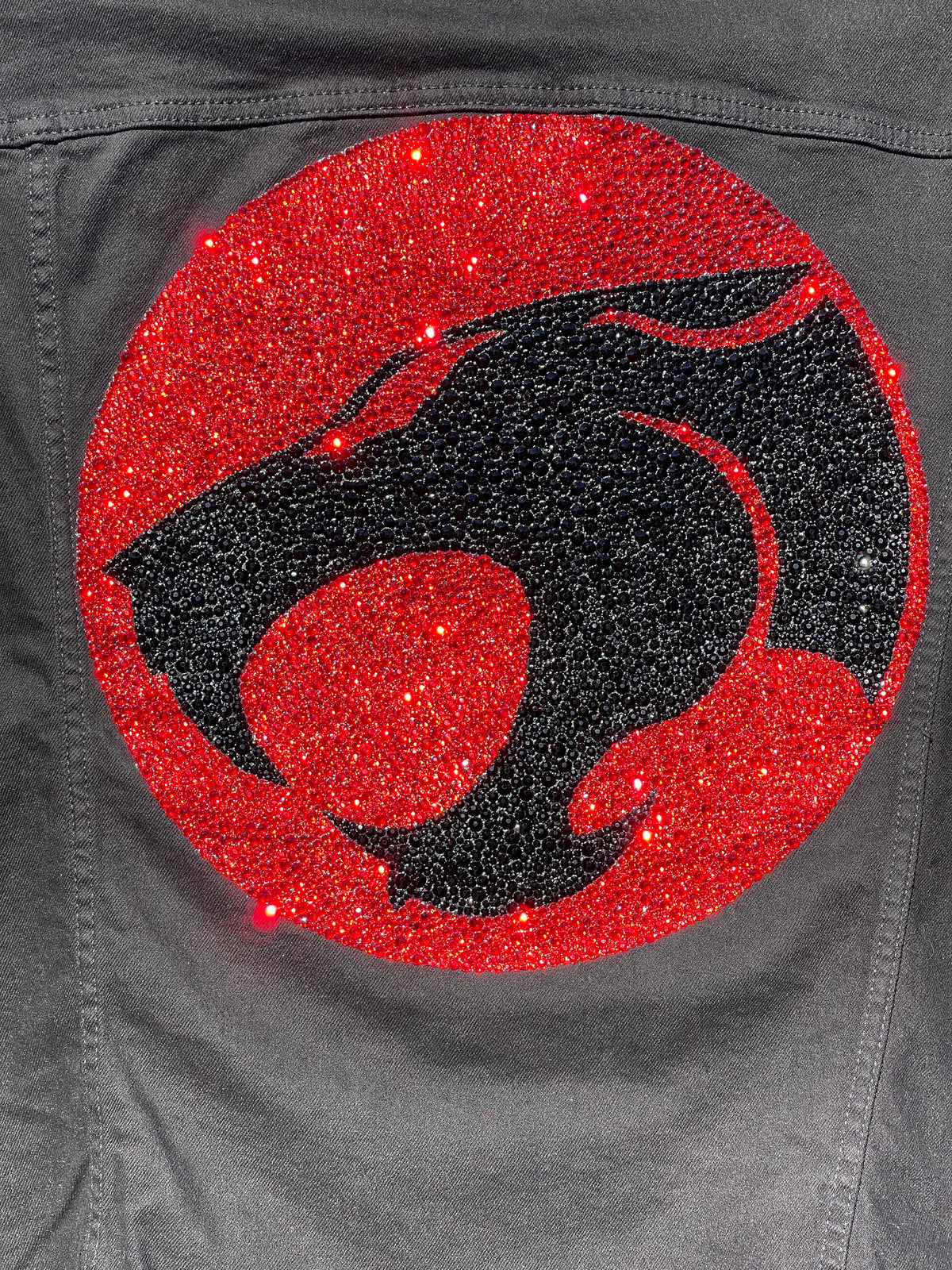 Black and red custom rhinestoned Thundercats emblem on a gray background. The grey background is a jean jacket