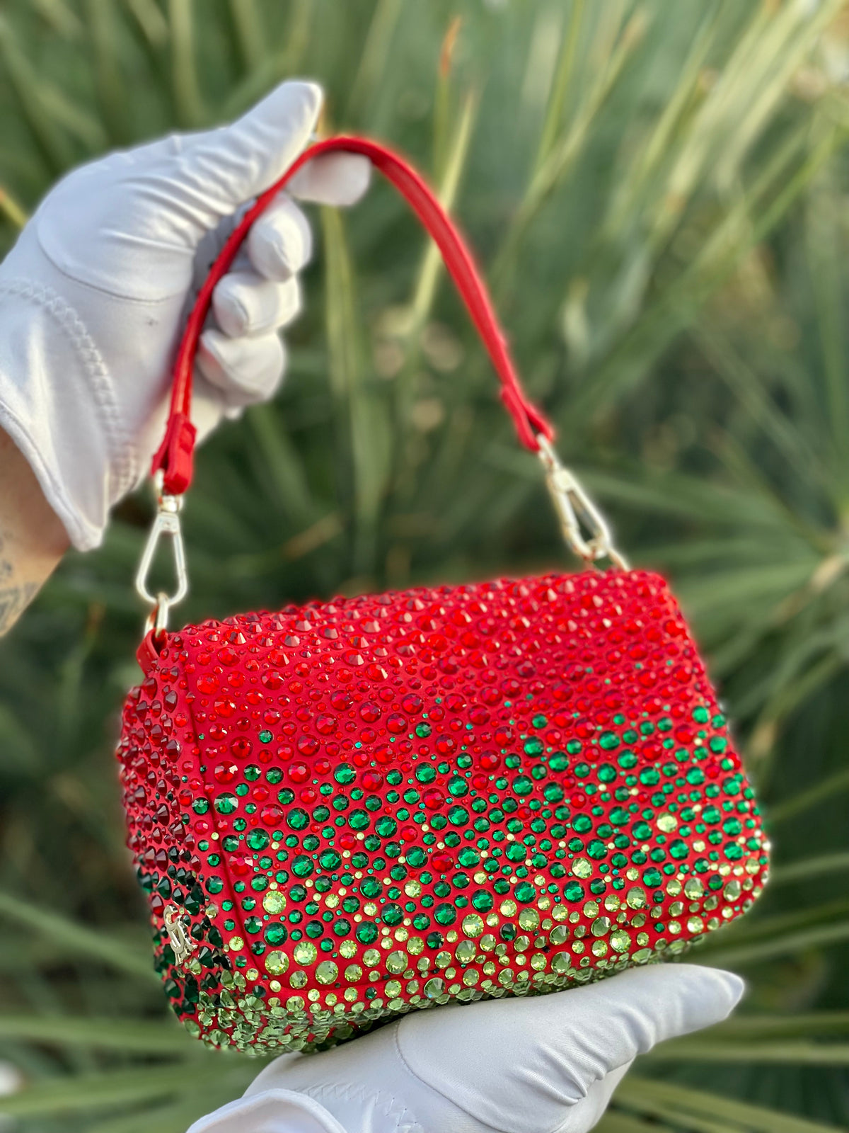 Christmas red custom rhinestone handbag with green and gold embellishments made by Looking Glass Gems held by a gloved hand against a blurred green background